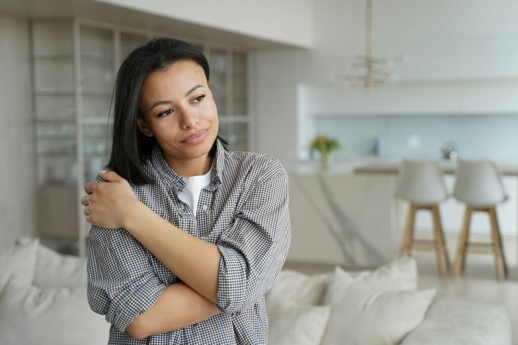 Calm happy female embraces herself feeling self love, self-pride, dreaming, standing at cozy home